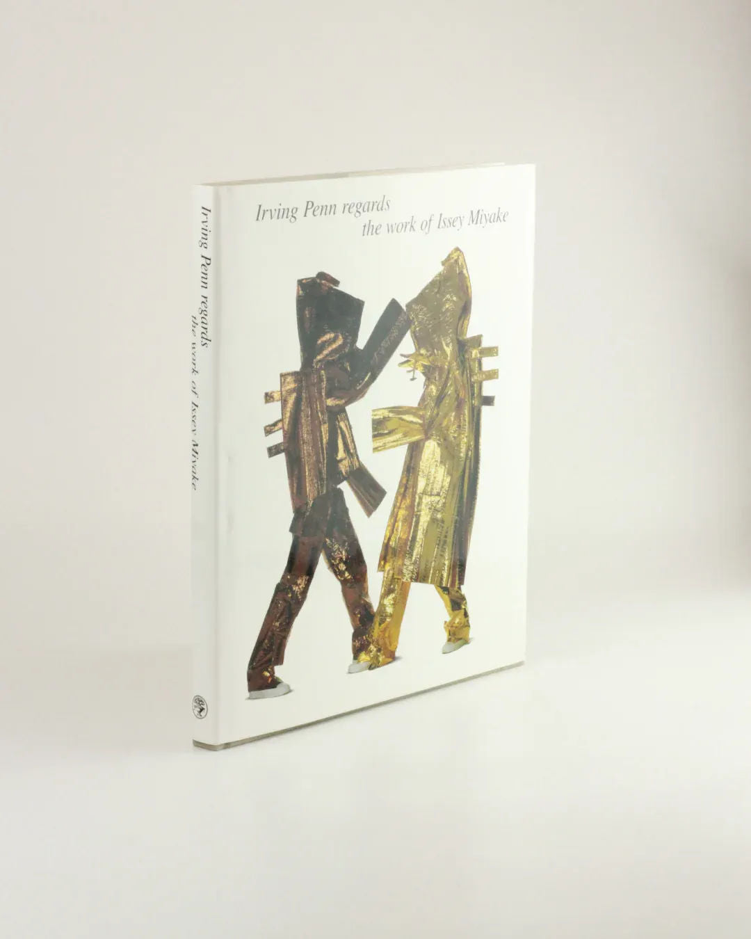 The image showcases the cover of the book "Irving Penn regards the work of Issey Miyake," available from Boga Avante Shop. The cover is characterized by a minimalist design that highlights fashion photography, featuring abstract metallic gold and copper figures facing each other in the center against a plain white background.