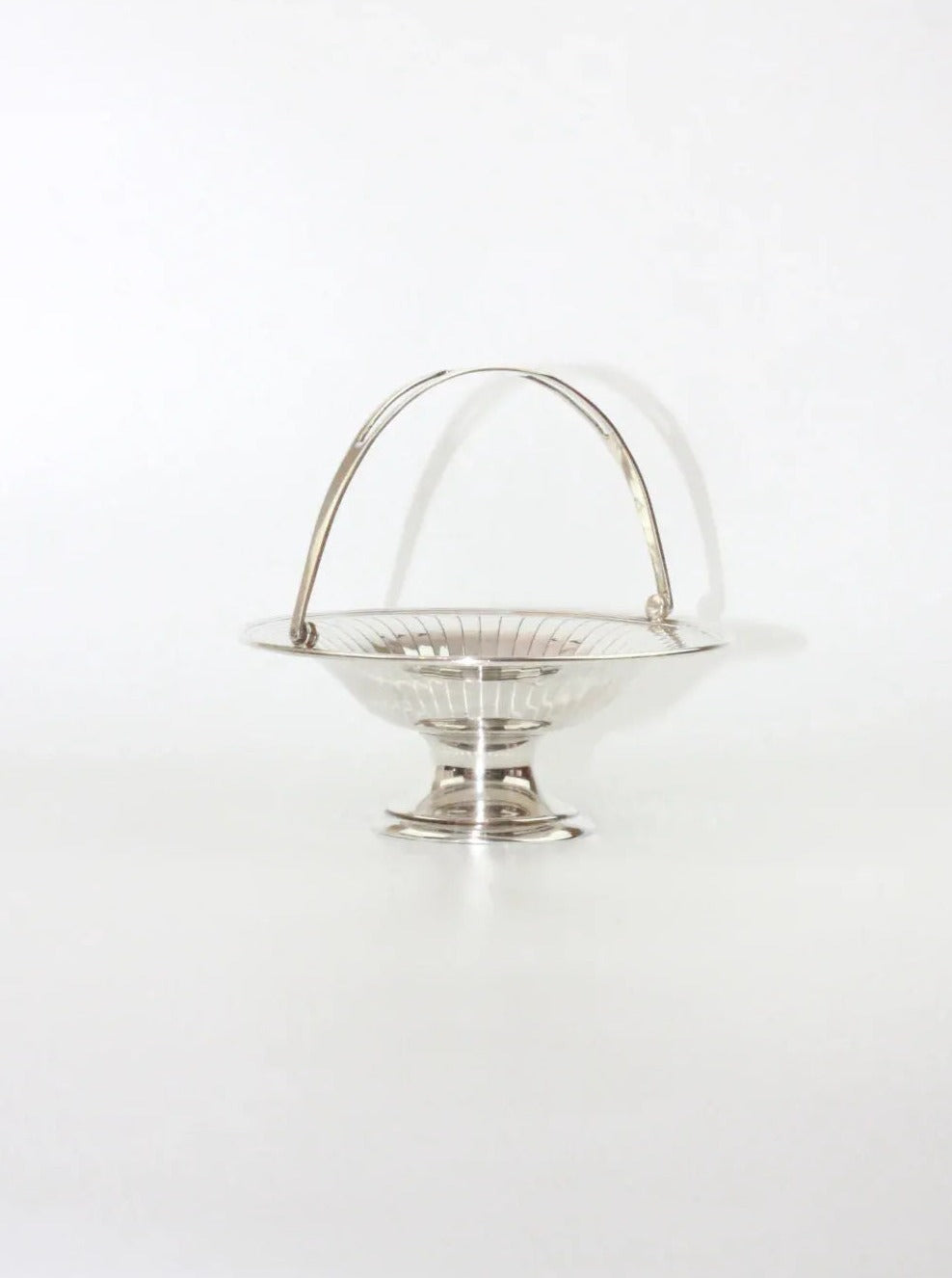The Silver Fruit Bowl from Boga Avante Shop features a nickel silver-plated basket with a polished, reflective surface set against a white background. The design includes an elegantly arching handle over its circular, shallow bowl, which rests on a small pedestal base. This vintage piece exudes classic, elegant appeal.