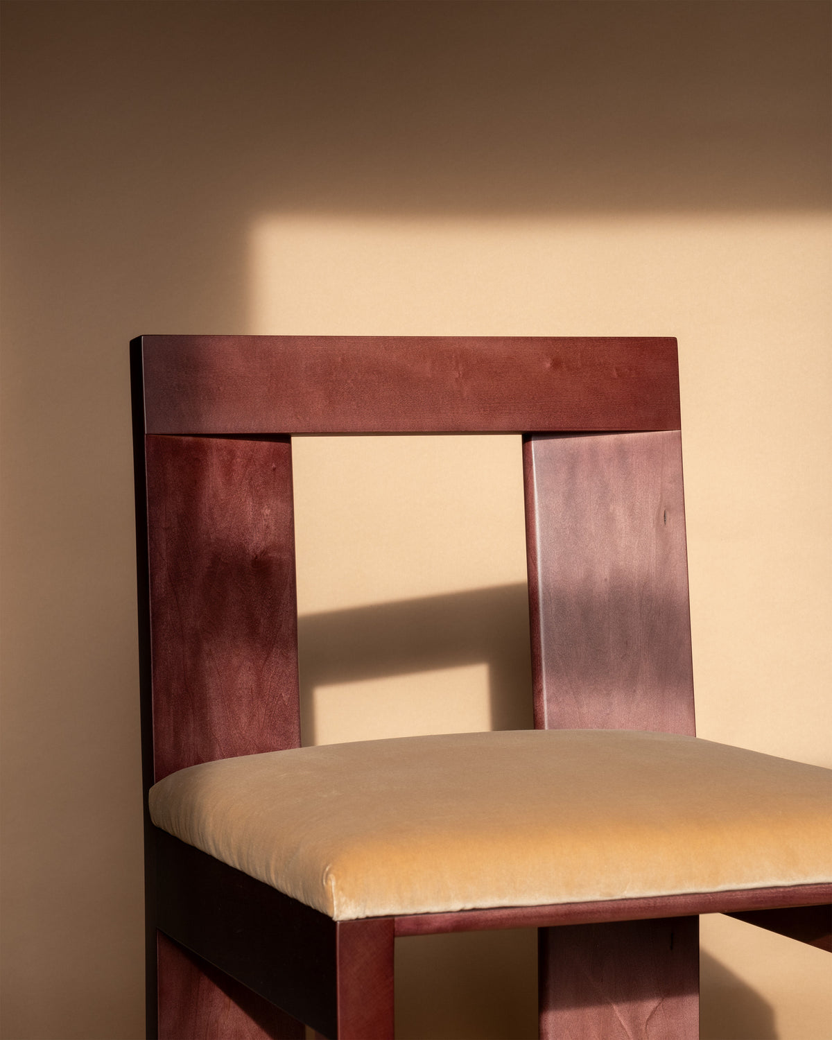 A Raja Chair by Juntos Projects, showcasing a minimalist design, is displayed against a beige backdrop. This contemporary furniture piece features a slightly curved backrest and an upholstered seat. The carefully arranged lighting casts the chair’s shadow onto the backdrop, highlighting the simplicity of the composition.
