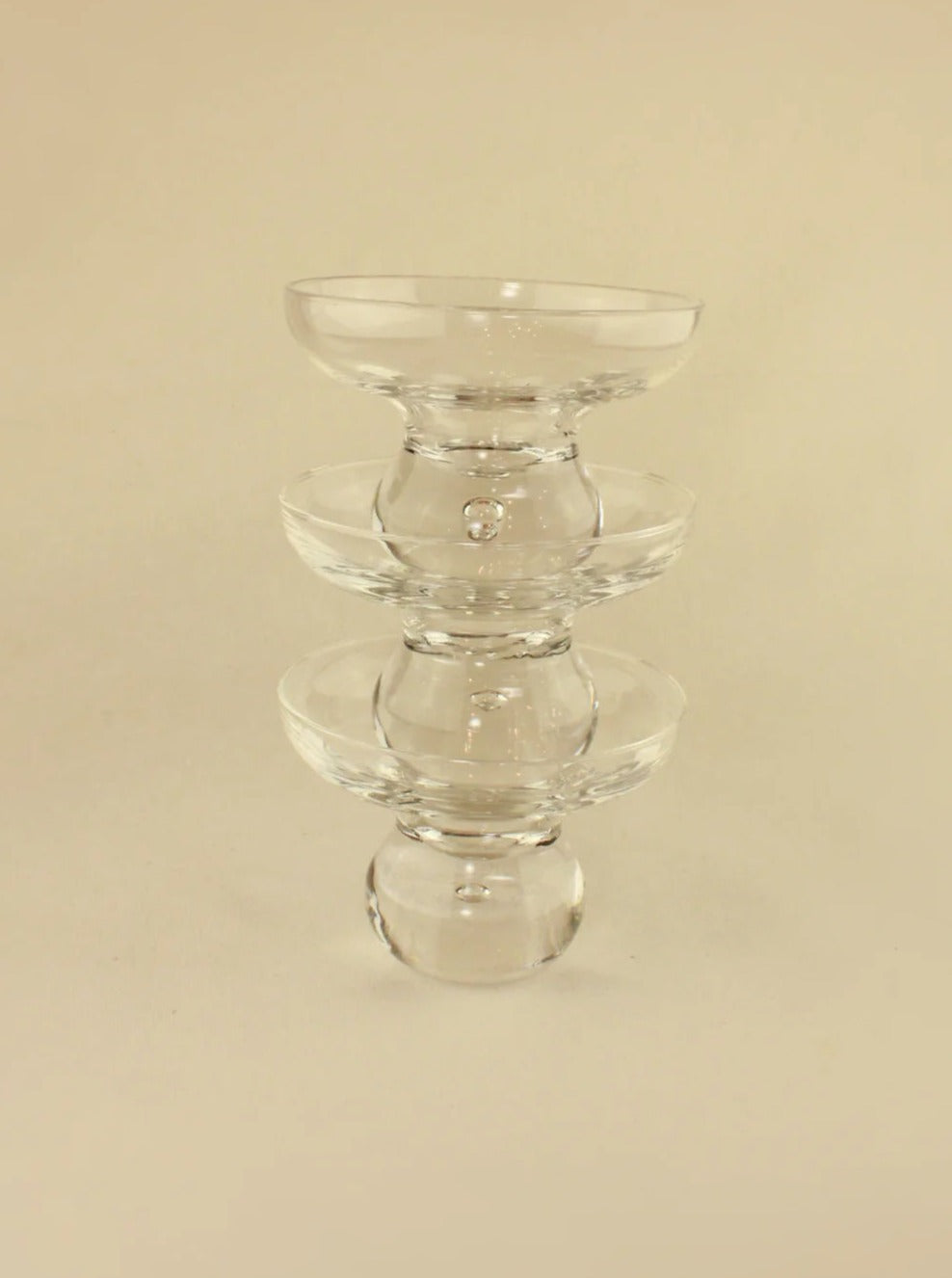 A clear glass sculpture featuring three stacked rounded bowls of decreasing size from bottom to top, reminiscent of vintage crystal glasses. The design is minimalist and symmetrical, standing against a neutral beige background. Introducing Italian Glasses by Boga Avante Shop.