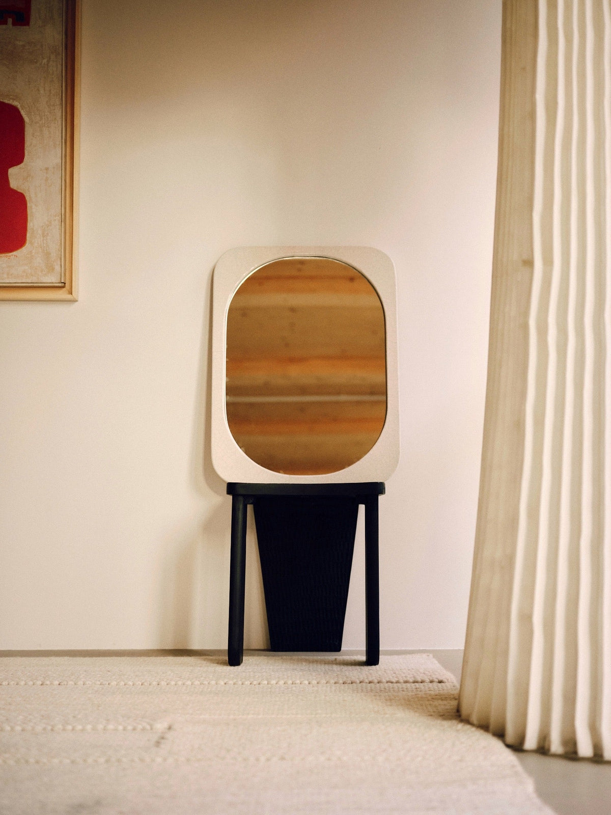 A small, rectangular, artisanal mirror with rounded corners— the Mirror Bien Fait in Lava Stone by Bien Paris— is mounted on the wall. The mirror is framed in a light-colored material and supported by a black stand. Made in Italy, it is positioned next to a beige curtain and a partially visible abstract painting with red accents.