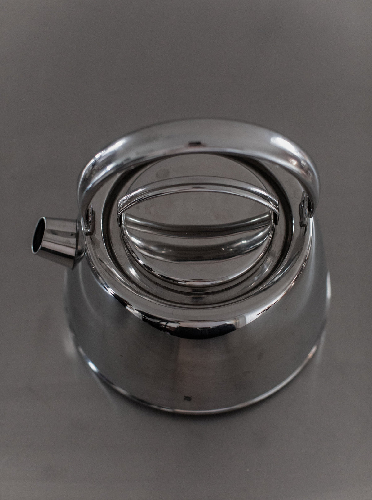 A polished Out For Lunch Chrome Teapot with a sleek, modern design is placed on a reflective gray surface. The teapot features a smooth, curved body, a short spout on the left, and a round lid with an elongated handle on top, enhancing any dining experience with its aesthetic appeal.