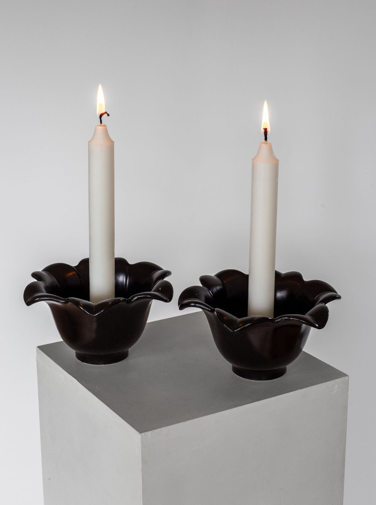 Two lit white candles stand in dark, flower-shaped holders made of ceramic. These Mid-Century Ceramic Candlesticks by Spigel from 1950s Sweden are placed side by side on a minimalist gray pedestal, set against a plain white background. The soft glow of the candle flames illuminates the scene.
