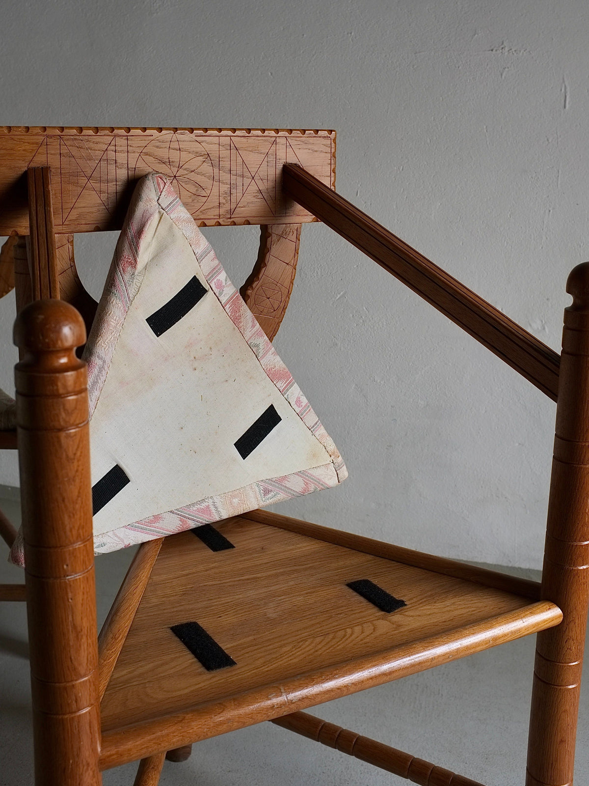 A uniquely designed Rustic Carved Oak Corner Chair from Veter Vintage, originating in Sweden in the 1950s. The chair, in vintage condition, features a detached seat leaning against the backrest, with four black strips, possibly Velcro, visible on both the cushion and the wooden frame. It is set against a plain, textured wall.