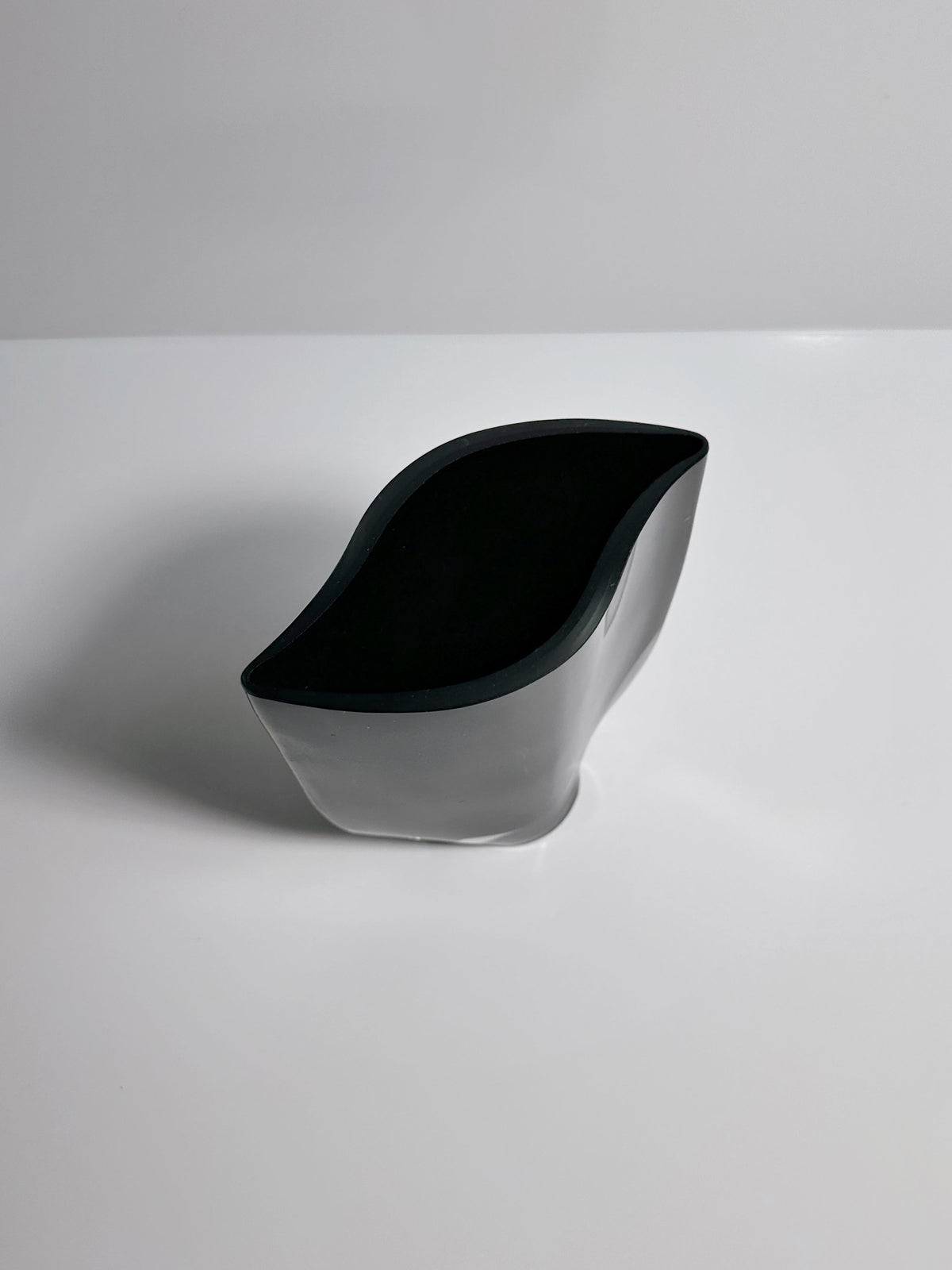 A sleek, abstract Black Glass Vase by Kaj Franck from Dodo Vintage is placed on a smooth, white surface. The vase features a modern, artistic appearance reminiscent of Finnish glassware, blending soft curves and angular lines to create an elegant and minimalist look.