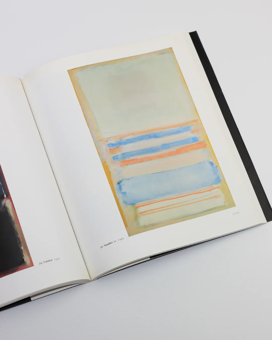 The Mark Rothko Book by Boga Avante Shop showcases abstract art akin to Colour Fields. The left page has dark artwork, and the right features light green, blue, and peach stripes. Both are titled "Untitled" and "Number 6," set against a background evoking Abstract Expressionism.