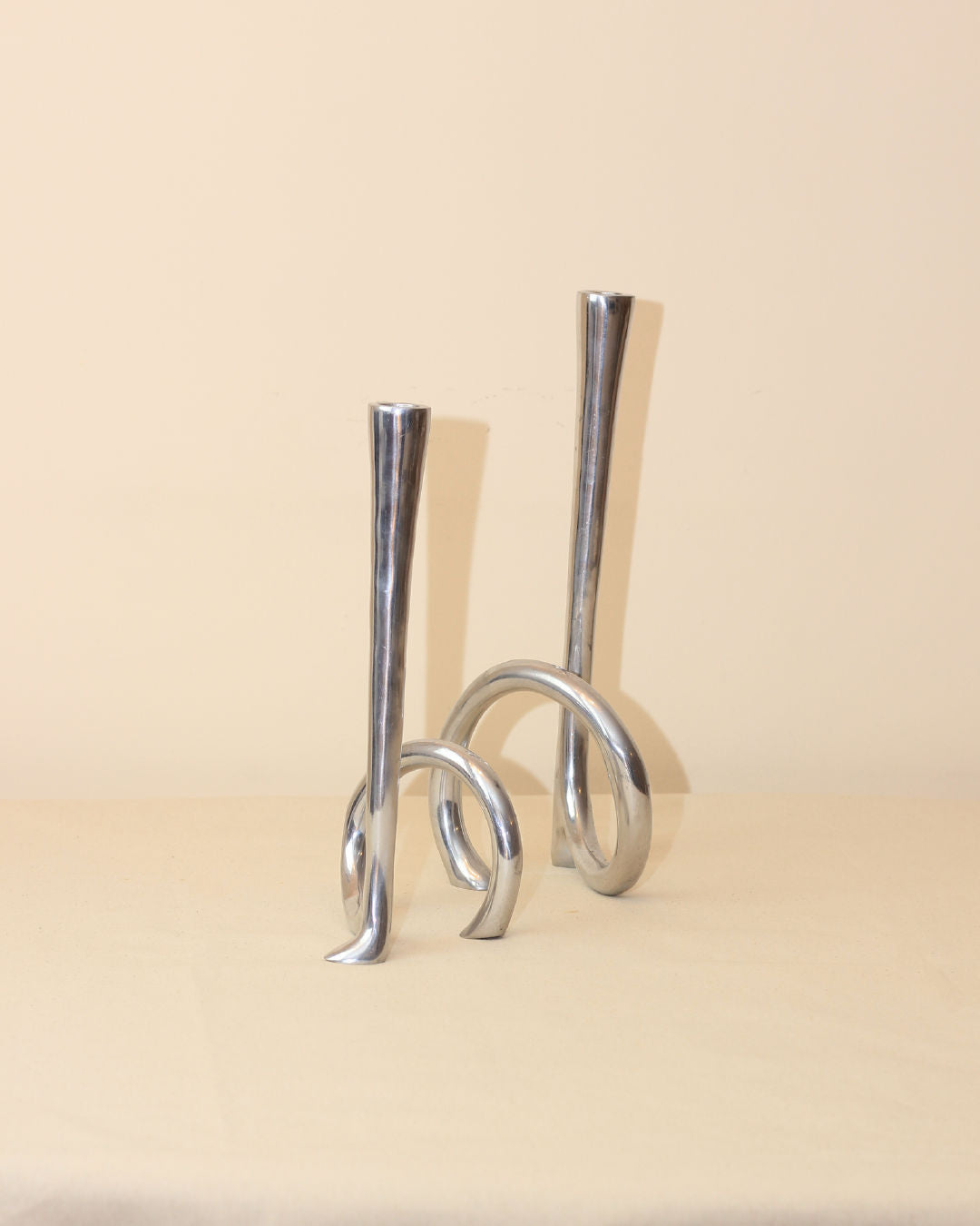 The Cast Aluminum Candleholders from Boga Avante Shop feature sculptural silver metal bases, shown on a beige surface and background—ideal for adding a modern minimalist accent to any space.