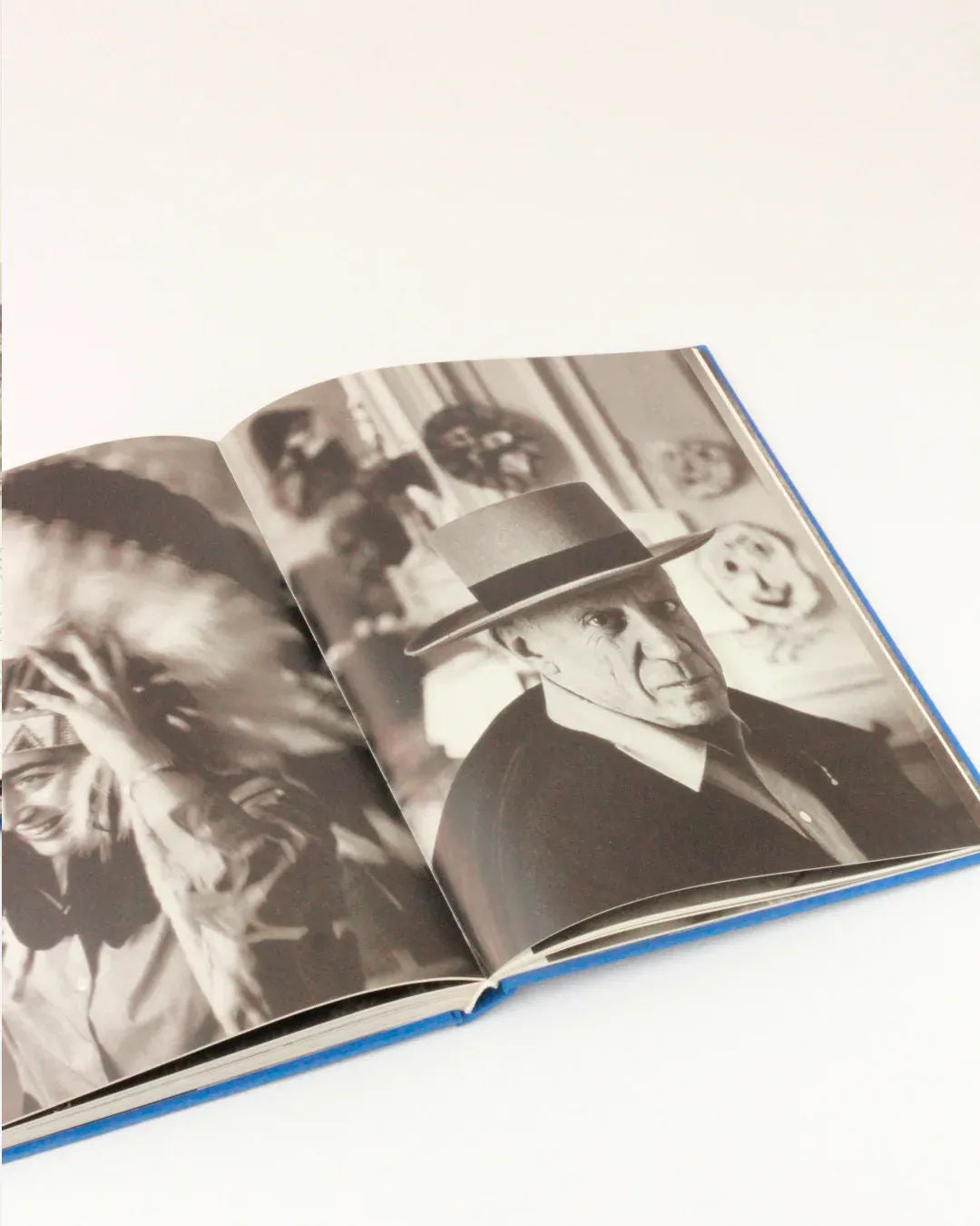 An open book with black-and-white photographs on its pages. On the left page is an intimate portrait of a person wearing a Native American headdress. The right page shows a striking image of Picasso and Jacqueline, staring ahead. The background appears to have other images or artworks from the Picasso and Jacqueline collection by Boga Avante Shop.