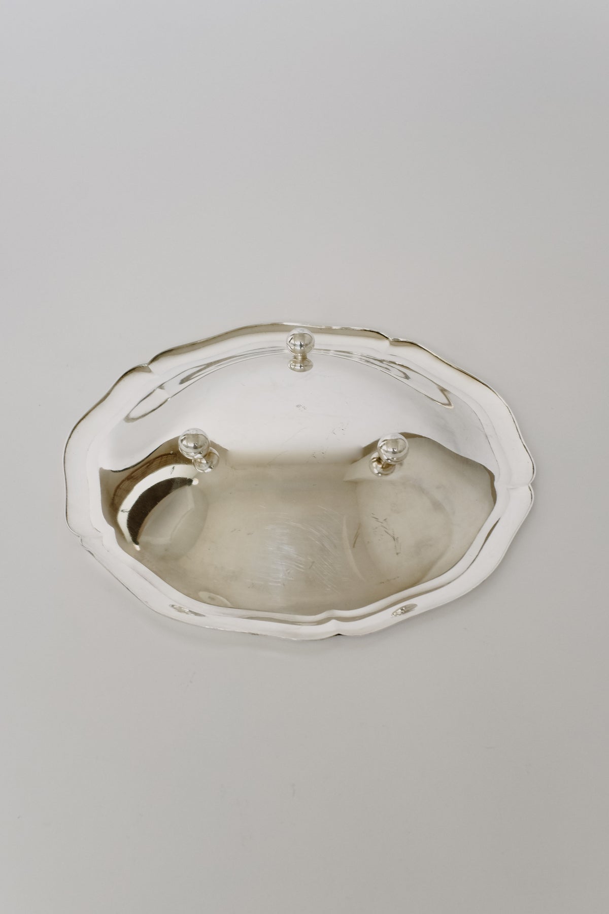 A polished Silverplated Bowl from septembre studios, featuring a scalloped edge and three small feet, rests on a plain white surface. This dish exudes vintage appeal with its reflective finish, showcasing subtle reflections and highlights.