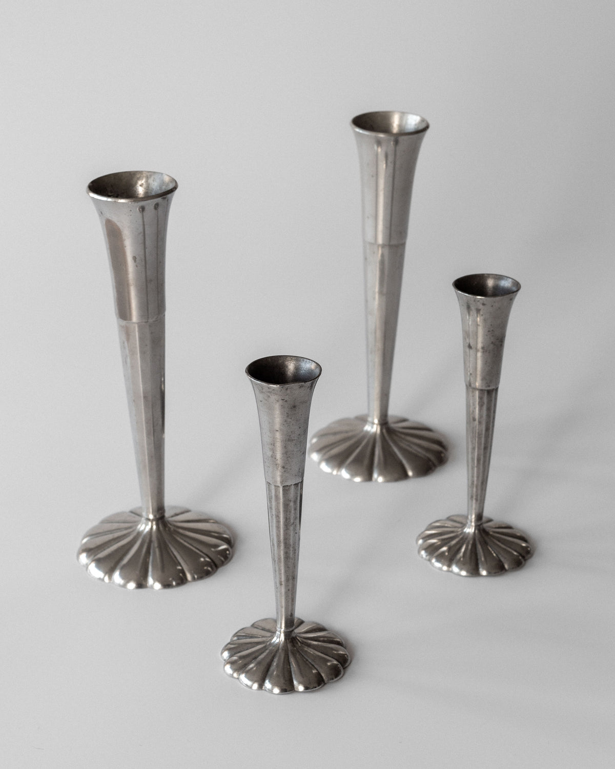 On a light gray surface are four varying-height vases from the Set of Four Art Deco Metal Vases by Spigel. Their silver, trumpet-shaped design with fluted bases and narrow stems embodies an elegant, minimalist Scandinavian aesthetic.