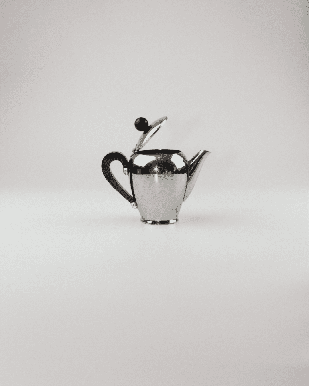 A sleek, modern silver teapot from the exclusive "Bombè" Alessi Tea/Coffee Set by Boga Avante Shop stands against a plain white background. The teapot features a rounded body, a black handle, and a matching black knob on the lid, which is slightly open.