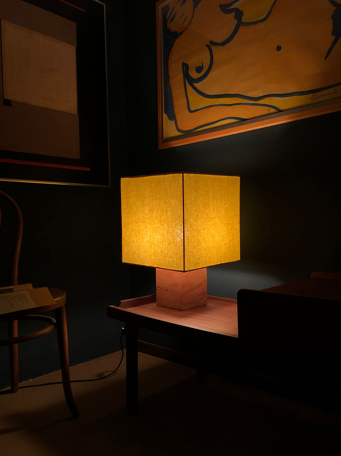 A softly lit room with a Roma I Squared Red Travertine from Nassi Lamps on a wooden base illuminating the space. The lamp stands on a wooden table next to a chair and a book. Artwork featuring abstract forms and a reclining figure adorns the dark wall behind, enhancing the ambiance with modernism and natural materials.