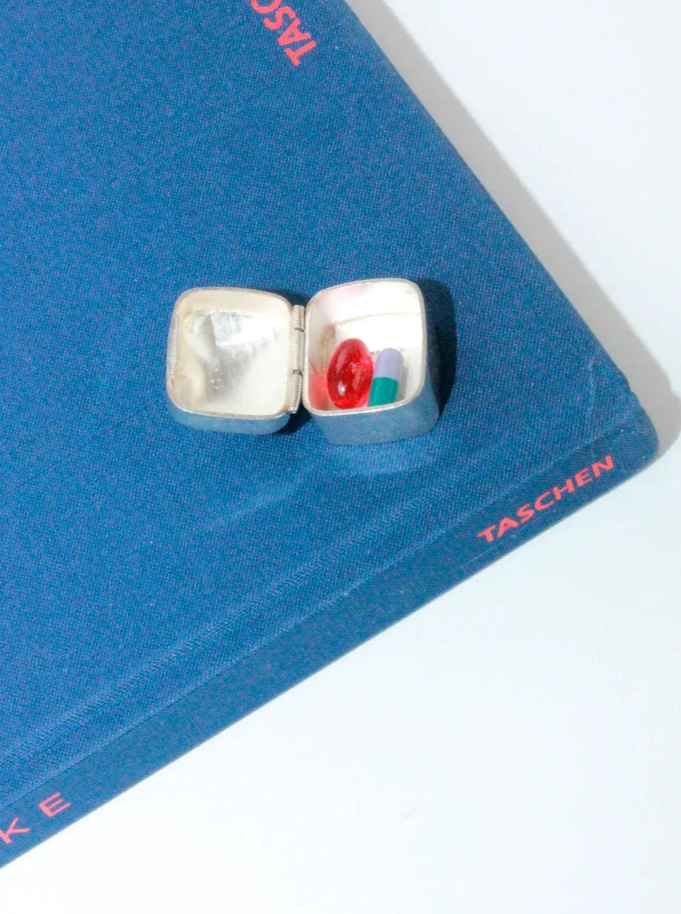 An open Silver Pillbox With Onyx Stone from Boga Avante Shop containing three pills—one red and round, one blue and long, and one white and capsule-shaped—is placed on top of a blue book titled "TASCHEN." The intricate design suggests it is handmade in Taxco, Mexico.