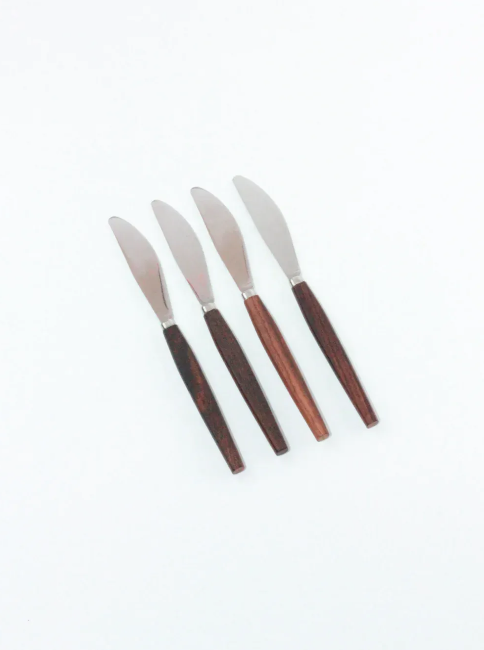 Four items from the Mid-Century Cutlery Set by Boga Avante Shop are evenly spaced on a plain white background. Each knife has a shiny stainless steel blade and dark teak handles, creating a simple and elegant appearance reminiscent of Mid-century cutlery. The blades are slightly curved and uniform in size.
