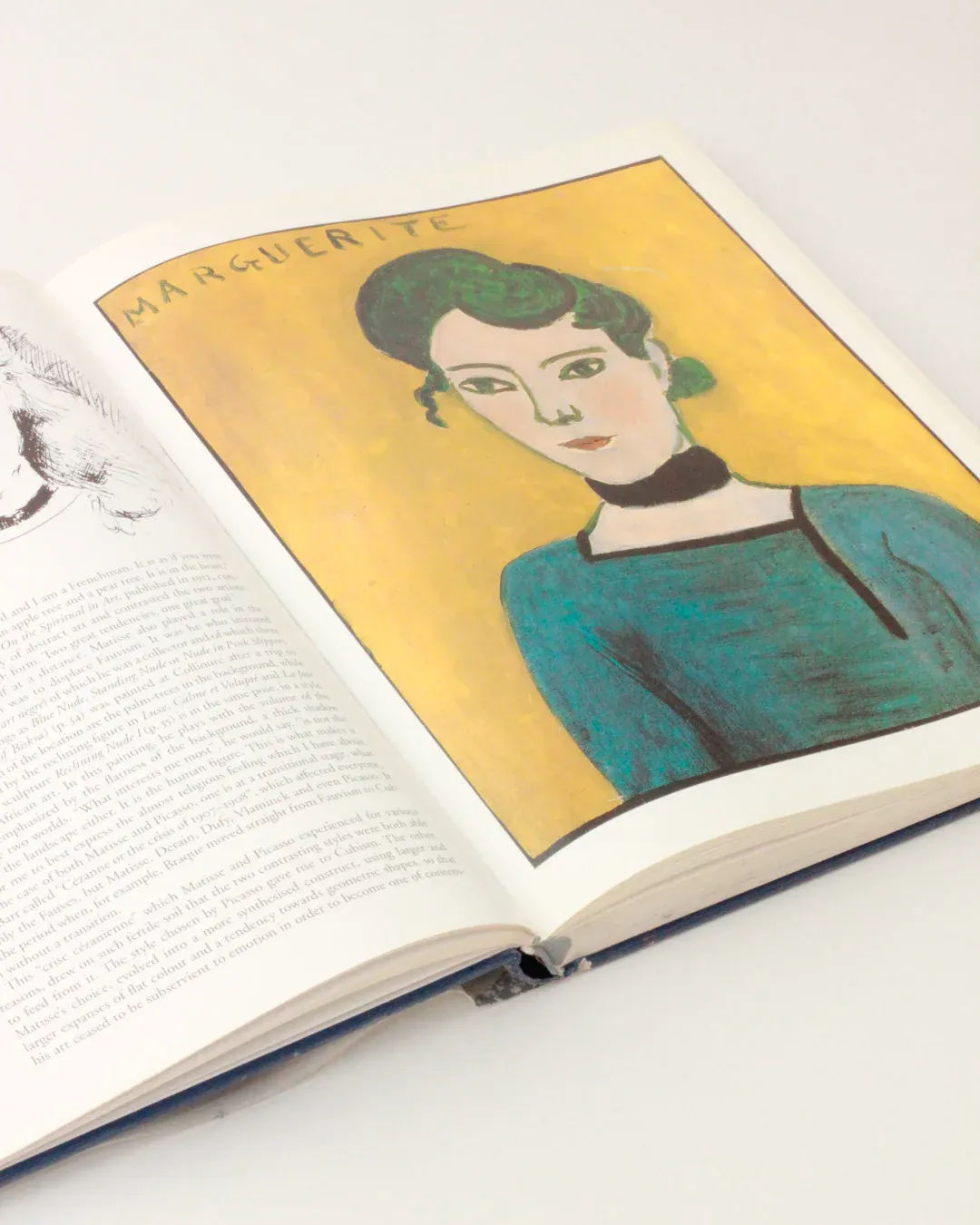 An open copy of the "Henri Matisse Vintage Book - Illustrated Publication" from Boga Avante Shop displays a colorful portrait of a woman with green hair and a green dress set against a yellow background, echoing Henri Matisse's style. The painting, titled "Marguerite," captures the essence of modern art. Printed text is found on the adjacent page. The book rests on a white surface.