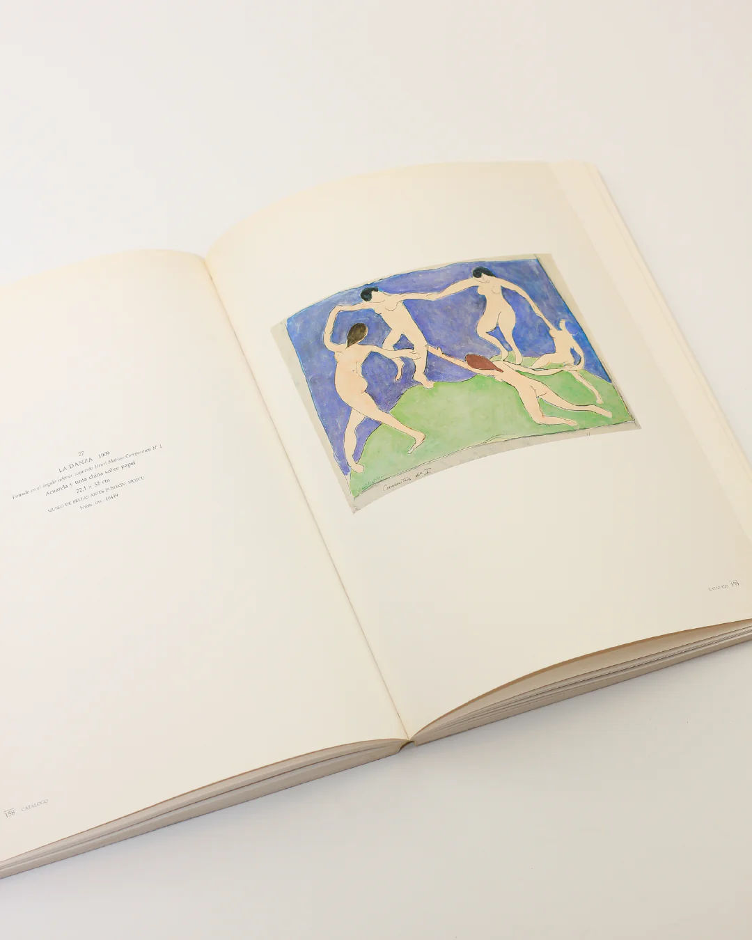 The Henri Matisse Book from Boga Avante Shop is opened on a light surface, revealing a painting by Matisse that depicts five nude figures dancing in a circle against vibrant blue and green backgrounds. This striking display of Fauvism is complemented by text present on the left page.