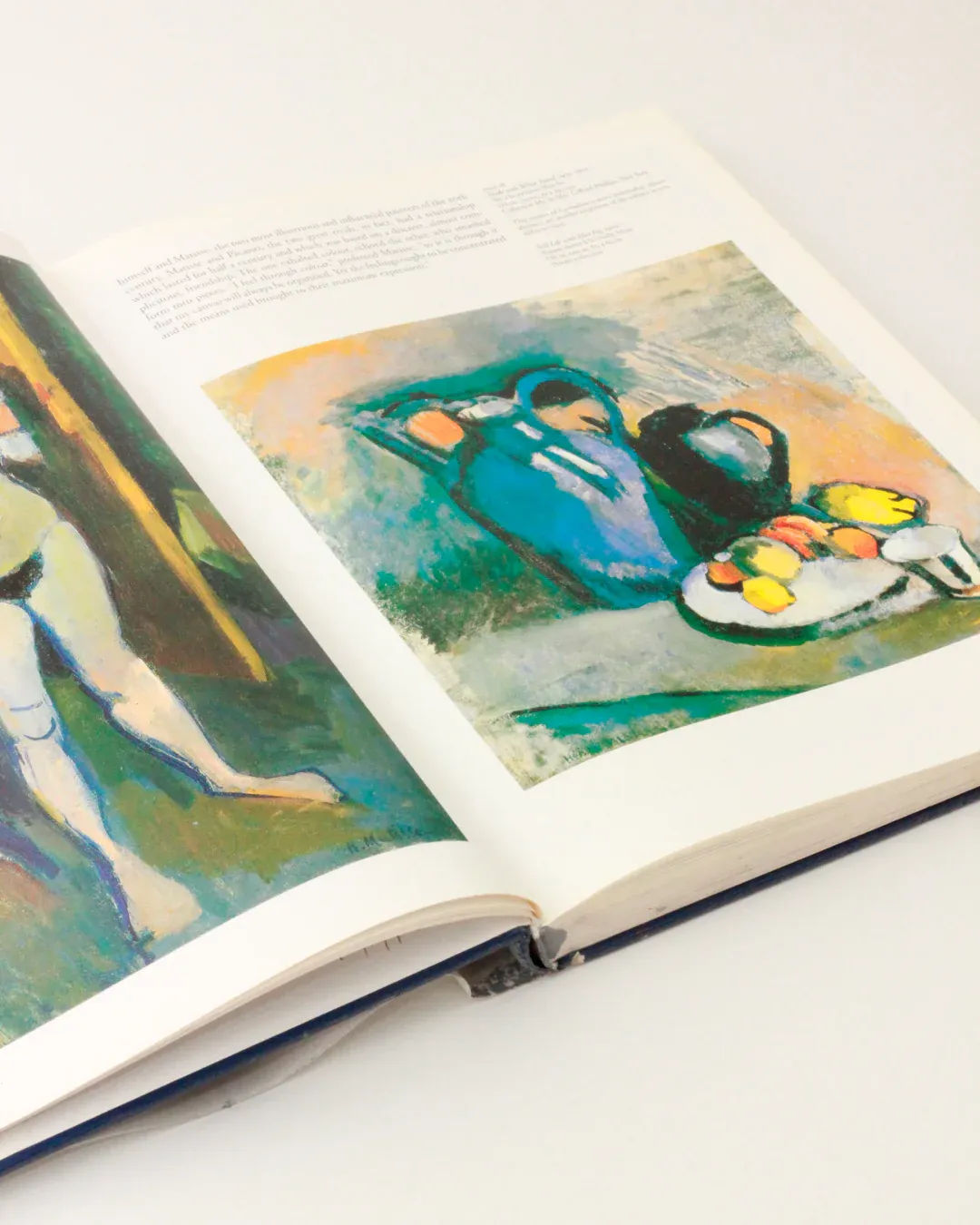 Displayed against a plain background is an open hardcover of the Henri Matisse Vintage Book - Illustrated Publication by Boga Avante Shop. The left page showcases a figurative painting with abstract elements, while the right page features a still life with jugs and a plate of fruit in vivid hues, capturing the essence of Matisse's modern art.