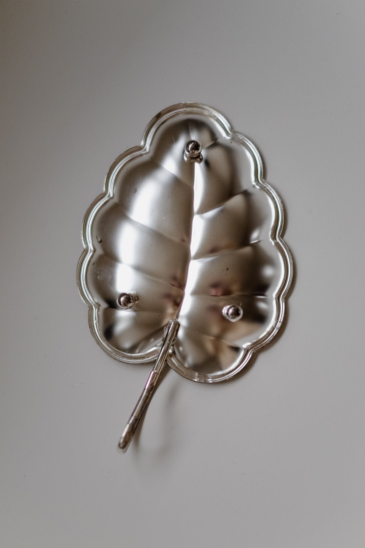 The silver "Serving Dish Leaf" by septembre studios, featuring a scalloped edge and small stem, elegantly rests on any surface. Its shiny finish reflects light, showcasing its textured design and infusing vintage appeal into your kitchen décor.