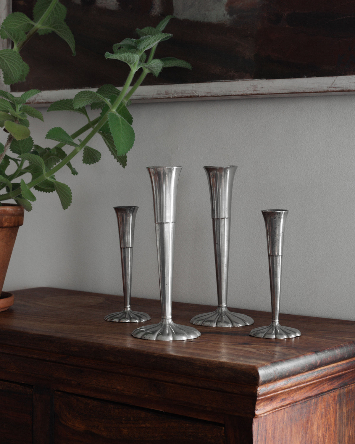 The Spigel Set of Four Art Deco Metal Vases, with their sleek metallic finish and varying heights, exudes elegance on a wooden surface. A leafy green plant in a terracotta pot provides a striking contrast against the neutral wall backdrop.