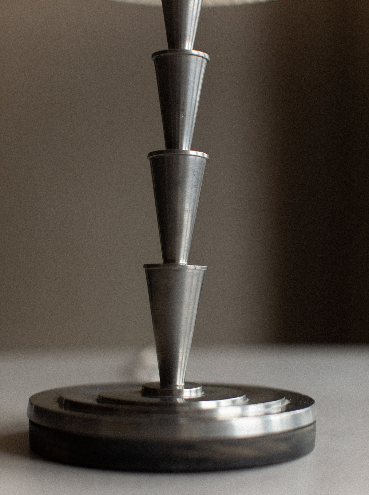 A close-up of a metallic lamp base, featuring a stack of four conical sections arranged vertically on a round base. The background is out of focus, emphasizing the sleek and modern design reminiscent of an Out For Lunch 1920's Swedish Grace Lamp by Elis Bergh.
