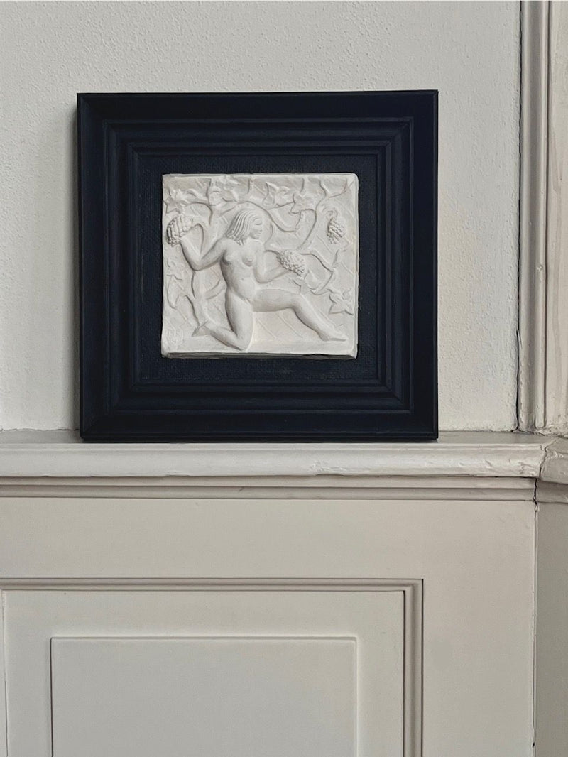 The Midcentury Plaster Relief by Bo Fjaestad, a Médecine product, features a reclining nude amid tropical garden vines. It's highlighted on a white, paneled wall by its black frame.