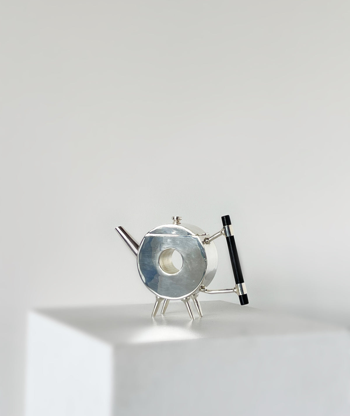 The Christopher Dresser Ornate Round Teapot by Porter Studio is a modern, circular silver-plated teapot with a black handle and angled legs, displayed on a white pedestal against a plain, light gray background.