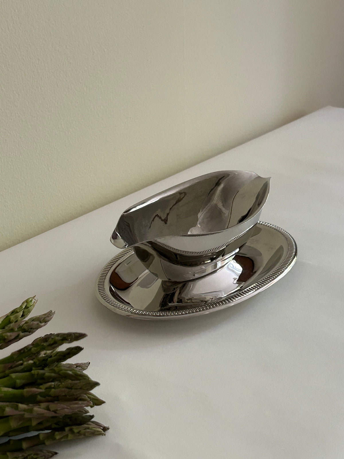 A beautifully crafted Stainless Steel Server Sauce from Flock Studio gleams atop its matching ornate tray on a white dining table, with fresh asparagus spears partially visible in the lower left corner.