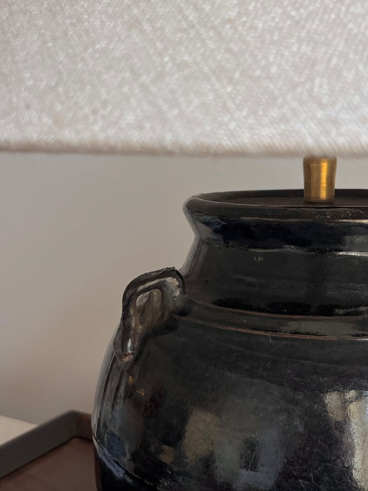 Close-up of the Médecine Antique Glazed Lamp with Ears, featuring a patina-coated ceramic base with a small handle and a beige fabric lampshade. The glossy base and textured shade enhance its vintage charm on the wooden surface.