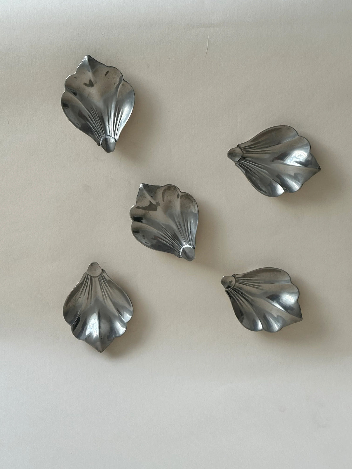 Five metallic objects resembling flower petals are neatly arranged on a light beige surface. Each "petal" has a textured, reflective surface and subtle grooves that mimic the natural lines of real flower petals, evoking the charm of Aesthete Label's Set 5 Vintage Chrome Leaf Trays.