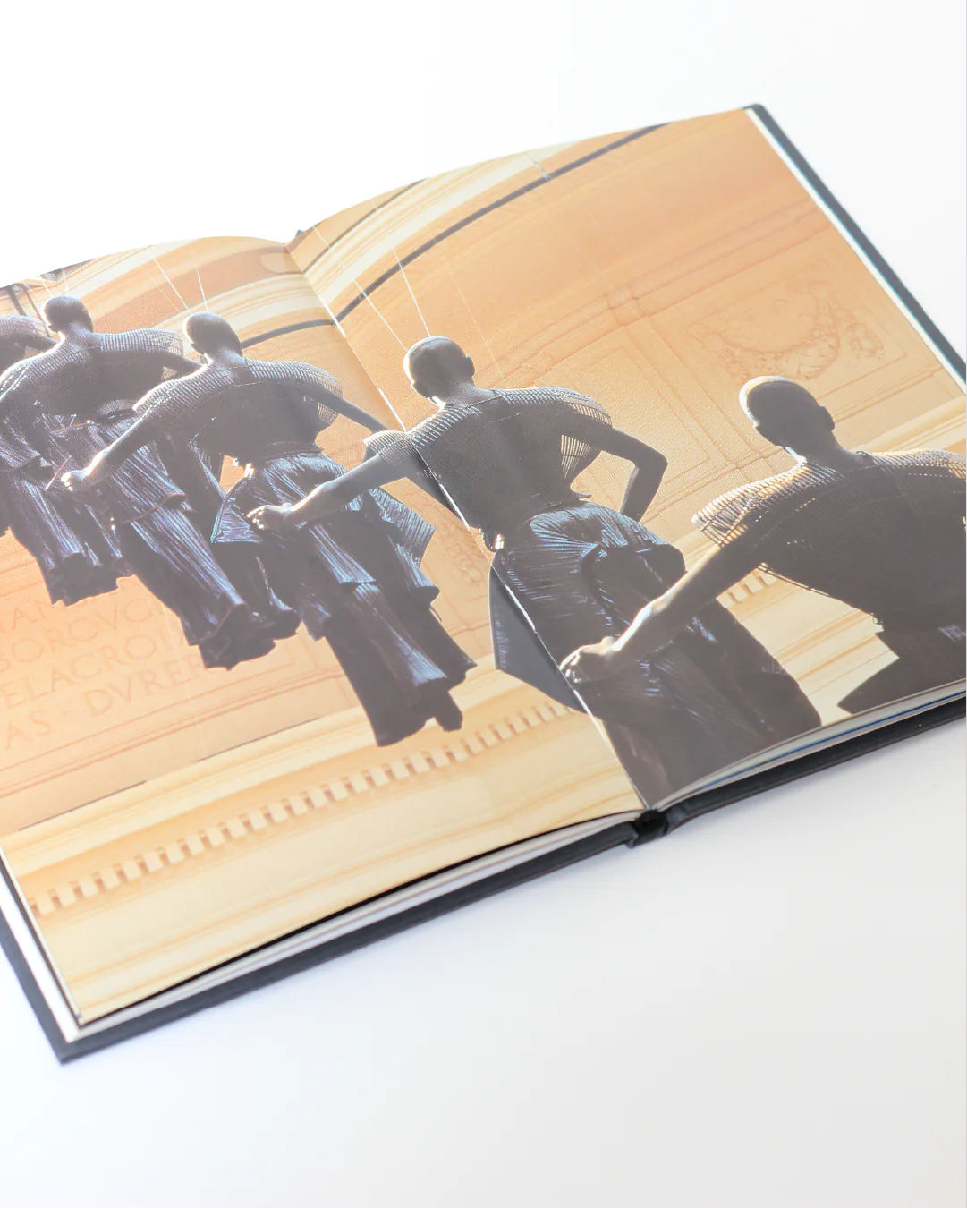 An open *Issey Miyake Book* from Boga Avante Shop reveals a photo of four dark-clothed, faceless figures with their arms extended forward, highlighting pleating techniques reminiscent of the iconic designer. Against a backdrop featuring a warm-toned architectural interior, this captivating image spreads across two pages.