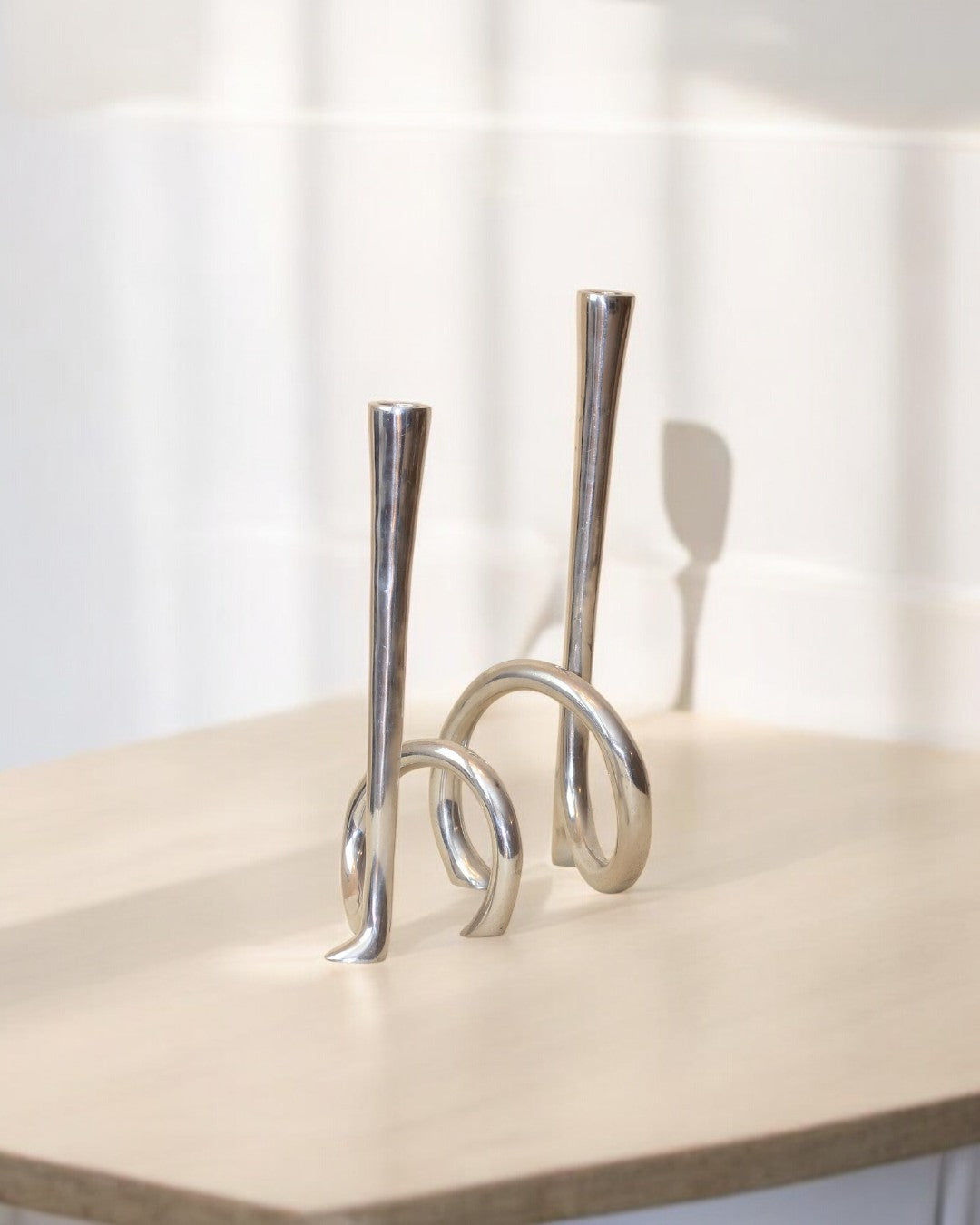 Two modern Cast Aluminum Candleholders by Boga Avante Shop, featuring curved, looped bases, sit on a light wood surface with soft shadows on a neutral background, recalling the sleek lines of 1980s candelabra.