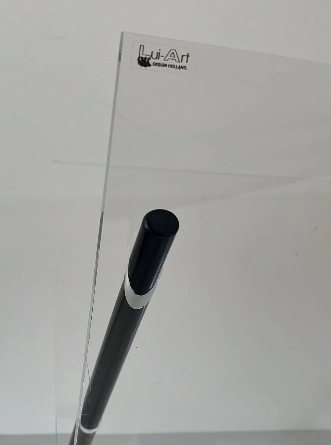 A close-up of a Magazine Holder Plexiglass by Lui Art Design with a black cylindrical object partially visible behind it. The top corner of the panel features a small logo that reads "CU-RA-TED." The background is plain and light-colored.