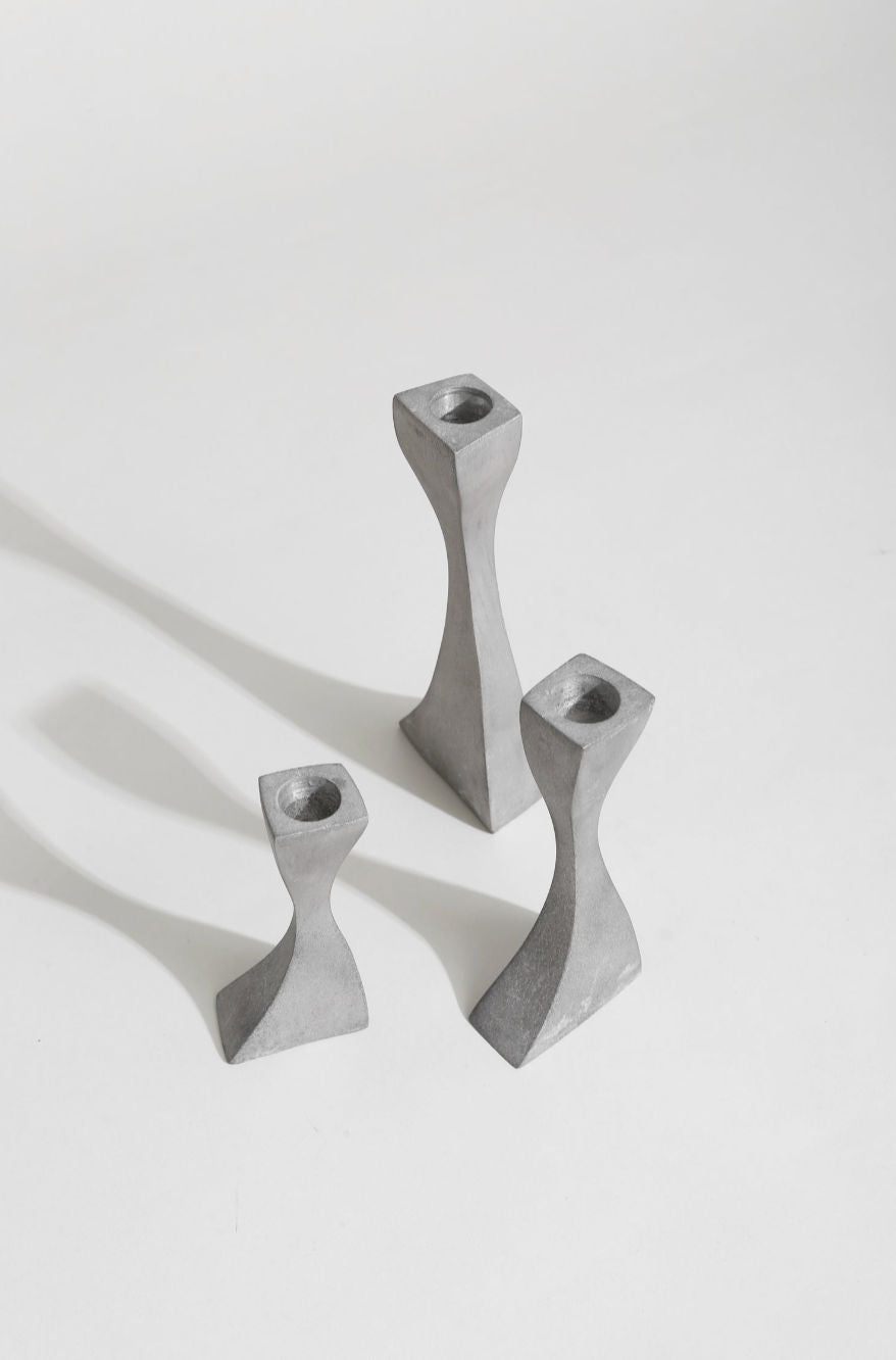 The Villa Wavy Candleholder Set features three modern gray concrete holders of varying heights and twisted geometric shapes on a white surface, casting soft shadows.