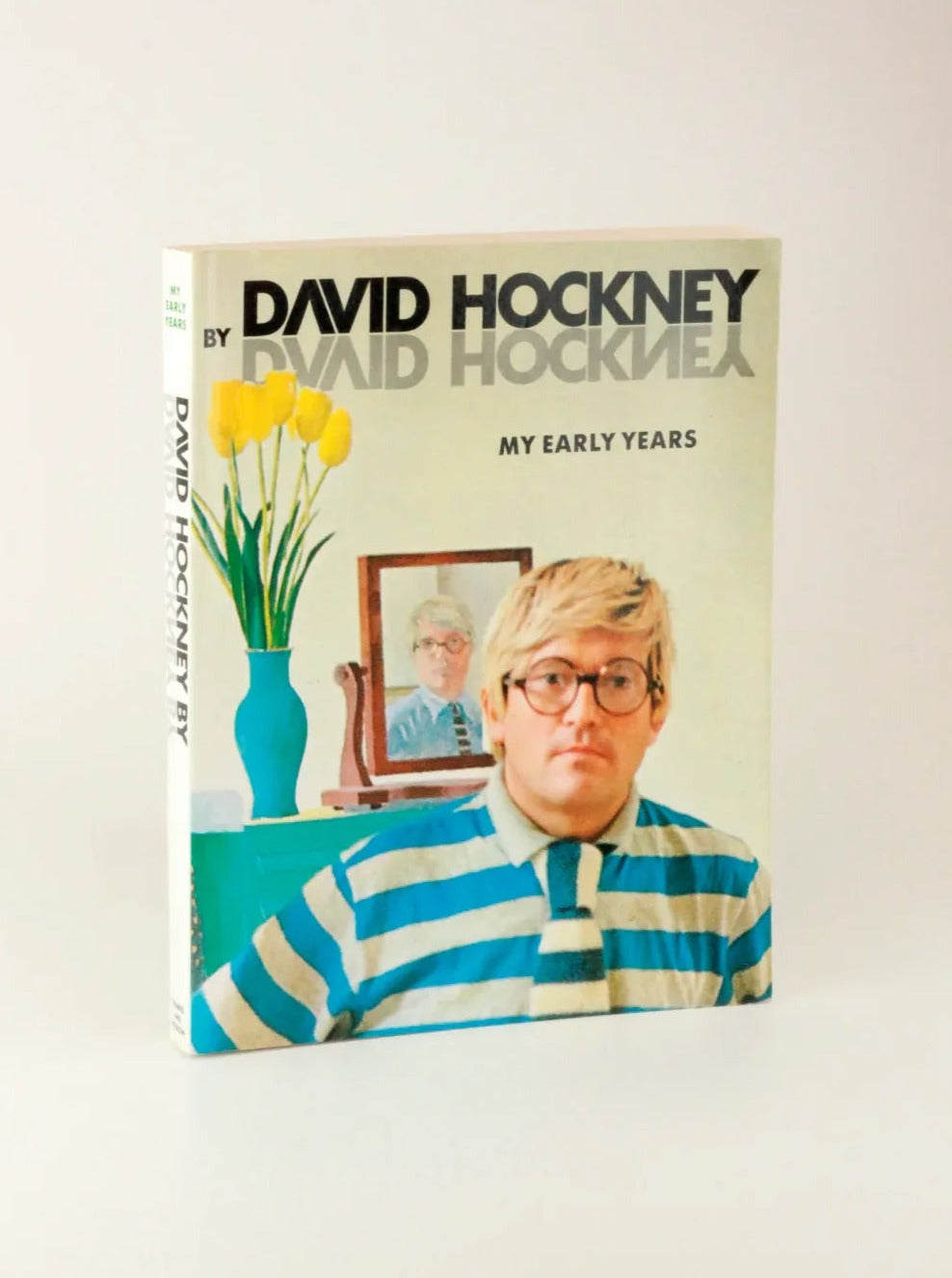 A libro vintage titled "David Hockney 'My Early Years'" from Boga Avante Shop. The cover features a photograph of the iconic figure in arte británico, wearing glasses and a striped shirt with a matching tie, standing in front of a mirror with a framed picture and a vase of yellow flowers beside him.