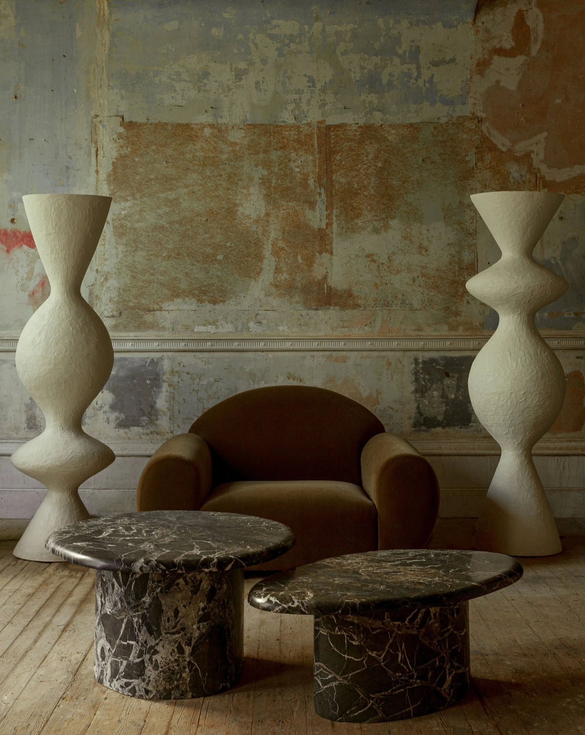 A rustic room features a "Sparta" armchair by Ransom & Dunn, elegantly draped in luxurious alpaca wool, and bordered by two tall, textured white sculptures. At the forefront, two black marble coffee tables with white veining sit upon a wooden floor. The background highlights an unfinished wall with earthy tones and textures.