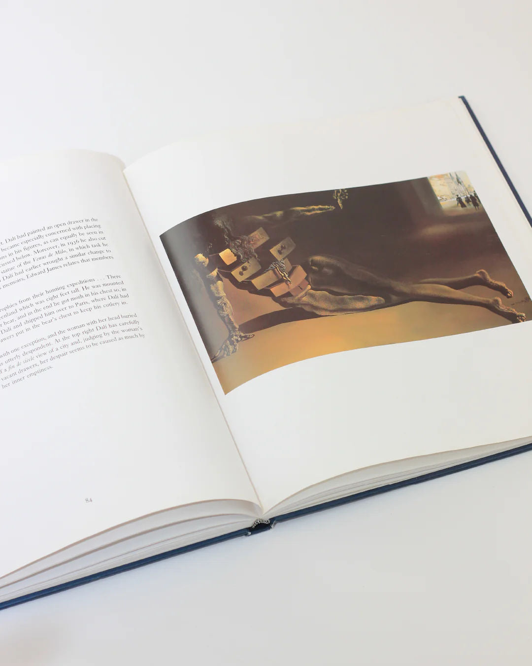 The Dali The Masterworks Book by Boga Avante Shop opens on a surreal painting reminiscent of Salvador Dalí's style, showing a distorted figure and abstract elements. The left page has text, while the book rests on a light surface, showcasing a minimalist, artistic layout.