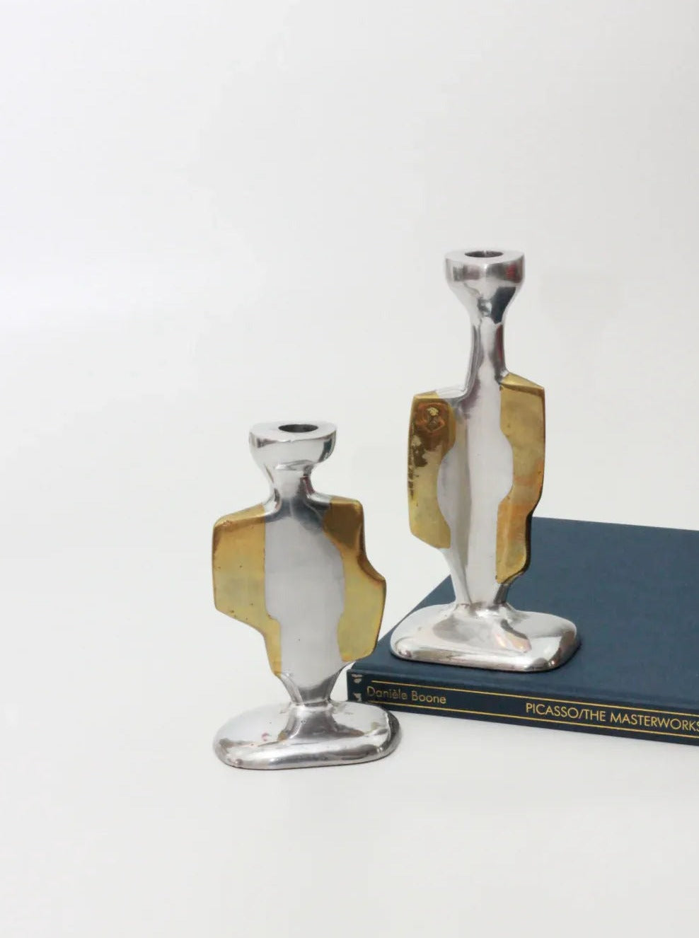 A pair of modern Brutalist Candleholders by Boga Avante Shop, crafted in silver and gold metal, evoke David Marshall's vintage style. They feature an abstract design with a gold accent in the middle and are elegantly placed on the book "PICASSO: THE MASTERWORK," which has a navy-blue cover with gold lettering.