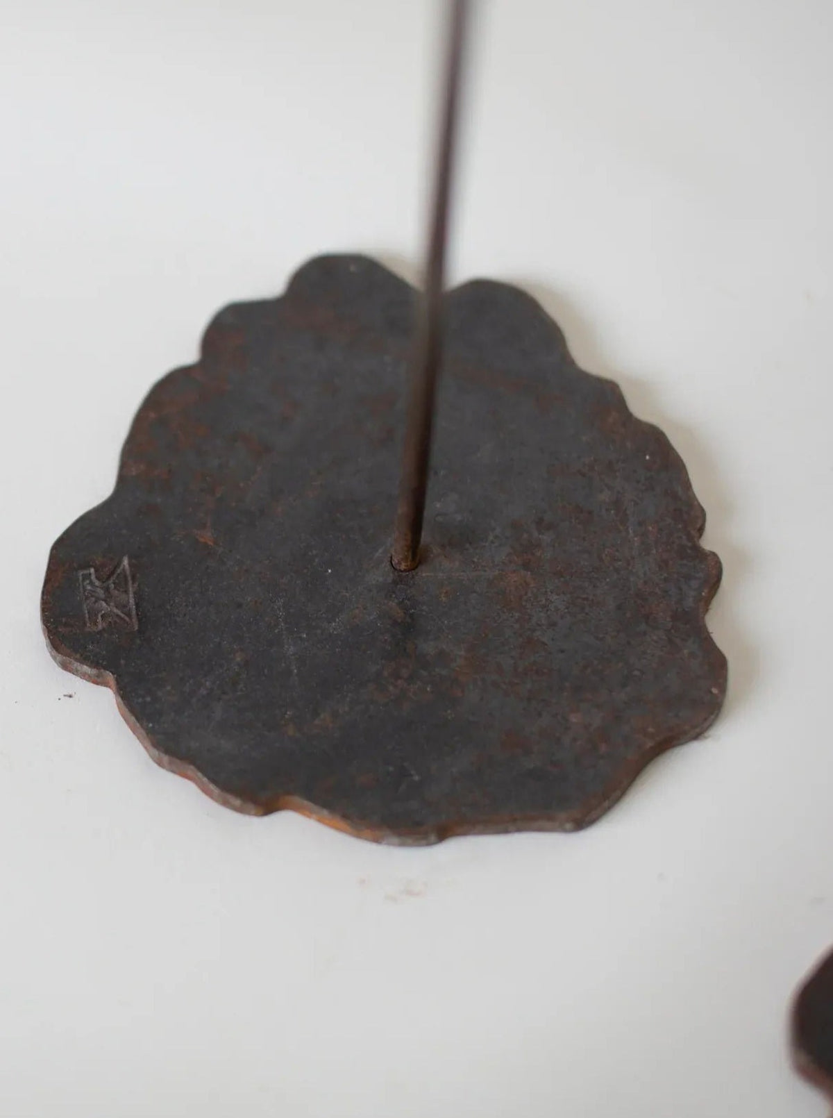 A close-up view of the Set of Copper Candle scones by Pieter Spruyt 1980s from CU-RA-TED, with a protruding stick for holding the incense. The base of the holder has an uneven edge and a small, engraved symbol on one side, reminiscent of handmade copper candle holders from the 80's, placed on a white surface.