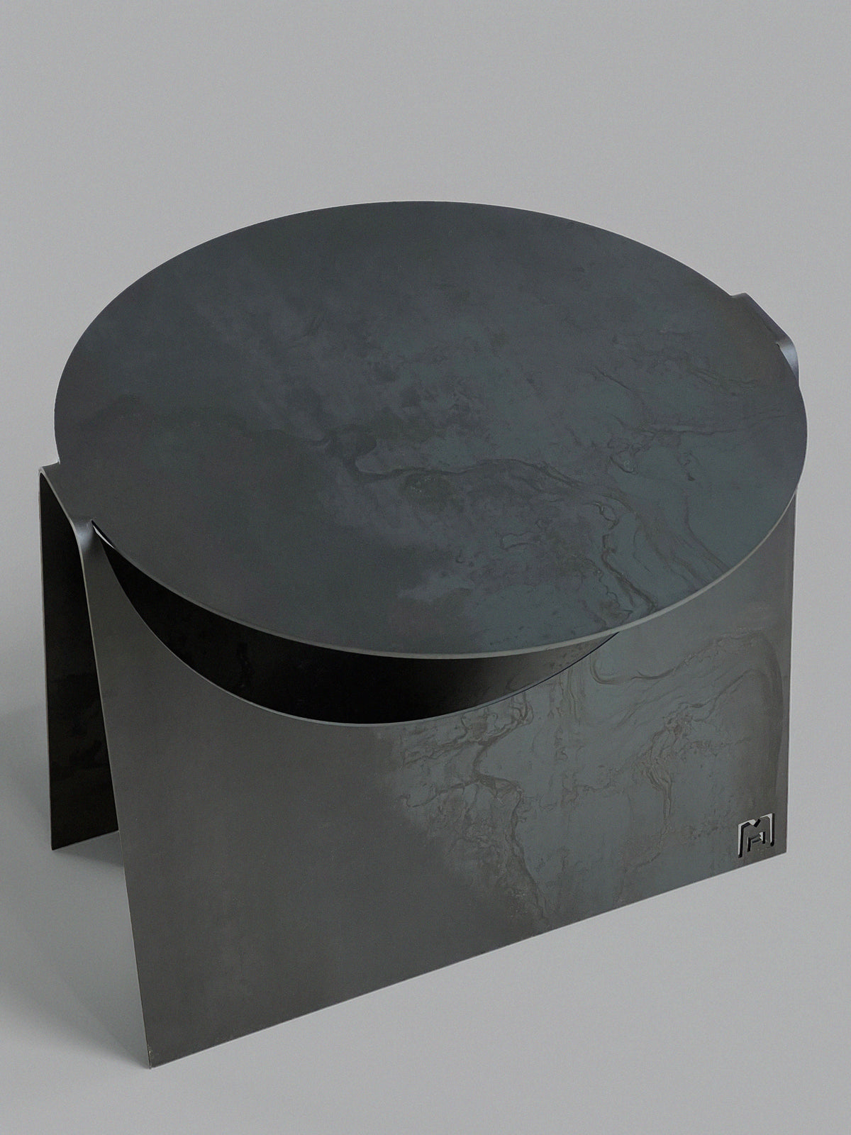 The 'Maru' Side Table by Modan features a modern, minimalist design with a round dark steel top, angular sides, a subtle textured finish, and a small geometric logo on one corner for distinct identity.