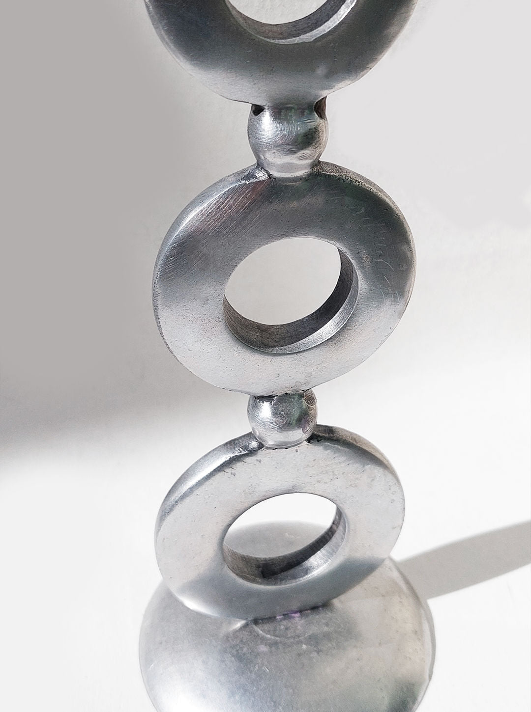 A close-up view of the Totem Candle Holder by Les Objoies reveals its classic shape, featuring three circular rings stacked vertically and connected by small spheres. The elegant silver plating gives the piece a refined look as it casts a soft shadow on a white surface.