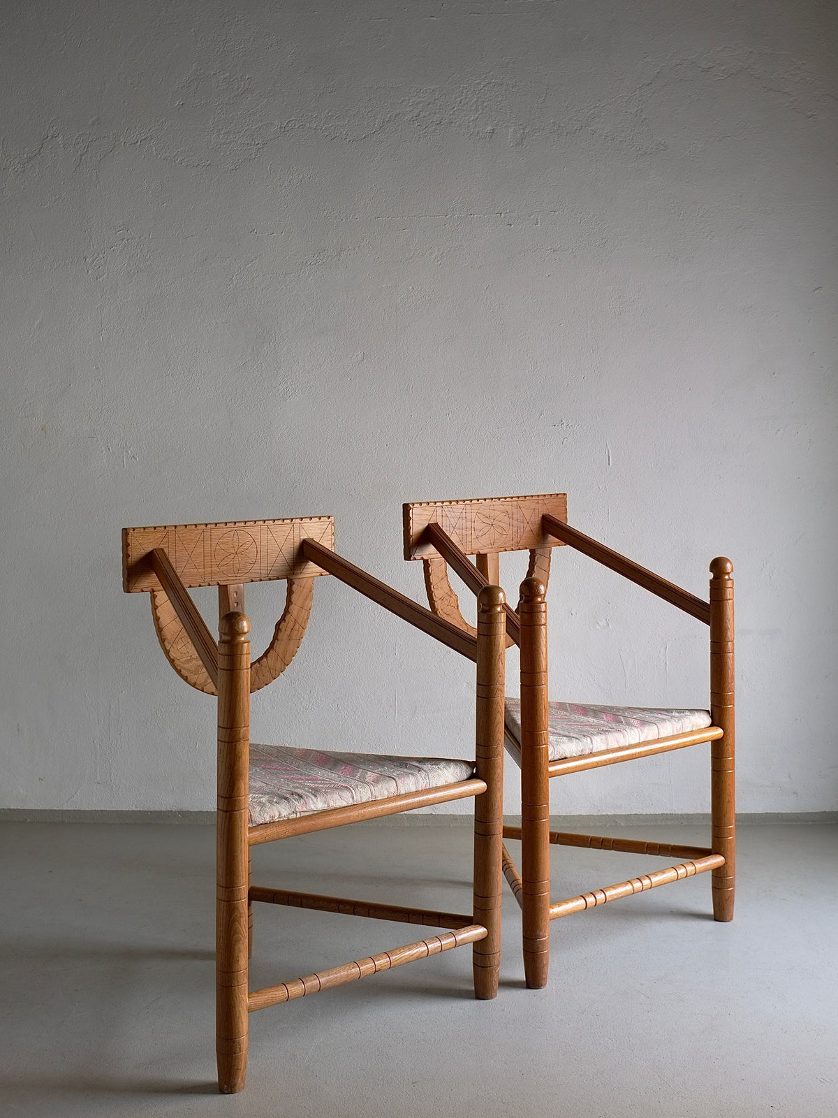 Two Rustic Carved Oak Corner Chairs from Veter Vintage stand side by side against a plain, light gray wall. Each chair from 1950s Sweden boasts a flat, rectangular backrest, a cushioned seat with a light-colored pattern, and simple, straight legs—classic elements of vintage design.