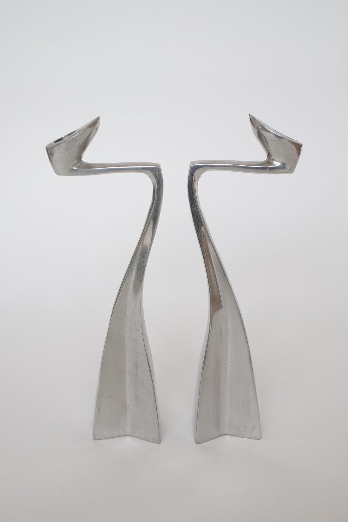 A sleek pair of Matthew Hilton 80's candleholders by septembre studios, featuring abstract twisted forms, stand side by side against a plain white background. Their curves and angles evoke dynamic movement and symmetry, making them exquisite collector's pieces.