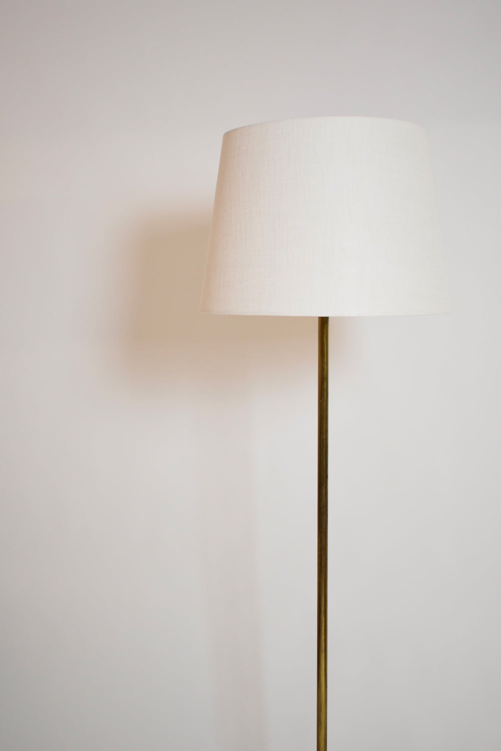 The Tripod Baluster Neoclassical Floor Lamp, 1960s by Barracuda Interiors features a slim brass stand and white lampshade, casting a soft shadow to the left against a plain white wall, evoking Riccardo Scarpa’s minimalist style.