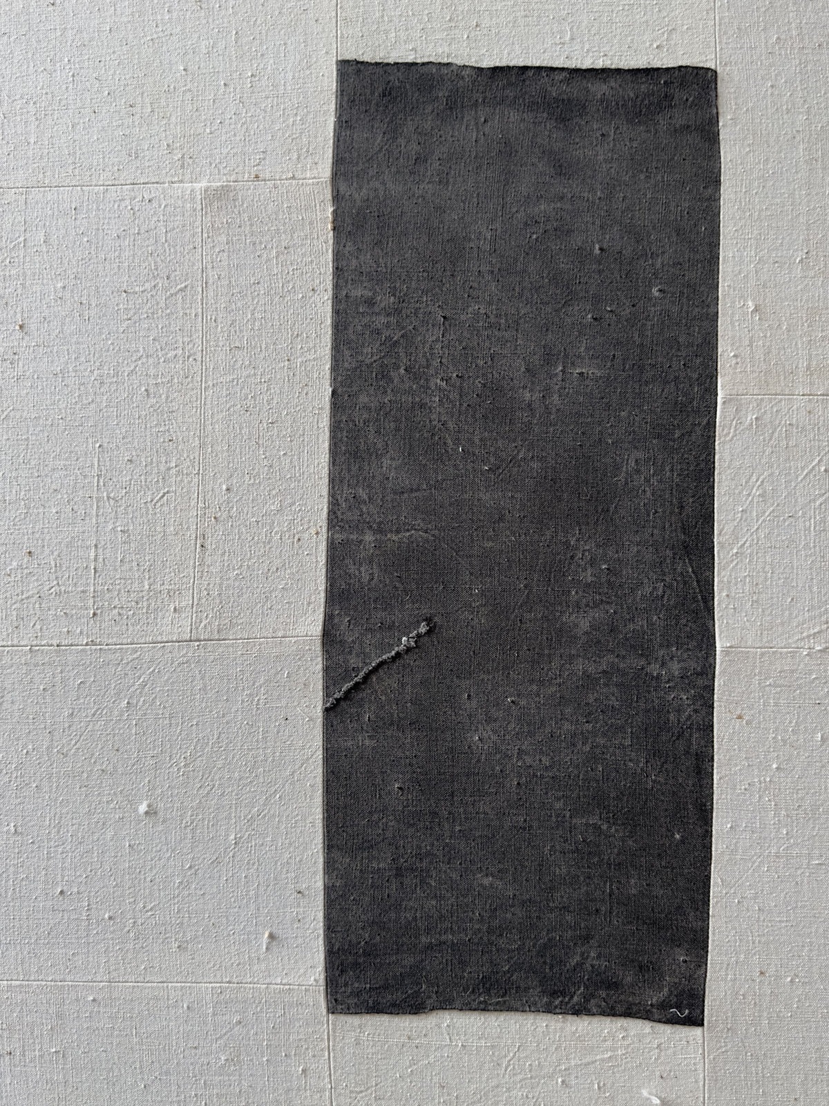 A black rectangular patch resembling a piece of Chinese cotton patchwork is adhered to a wall consisting of light grey square tiles. The (a-tent), 2024 Framed Hand Loomed Tapestry by Bec Kirby has a slightly textured surface with visible signs of wear and some minor tears. The tiles have a subtle, uneven texture, creating a contrast with the tapestry.