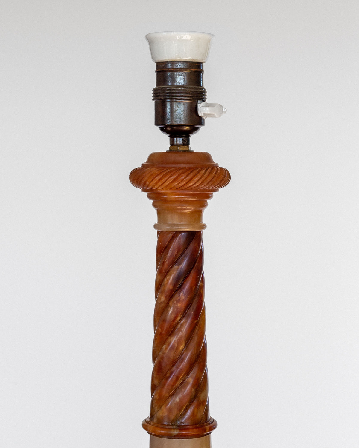 Introducing the Antique Alabaster Table Lamp, C. 1900 by Spigel—a vintage lamp base showcasing a twisted wooden column design with a bronze-colored socket at the top. While the light bulb and lampshade are absent, this antique table lamp exudes a classic and timeless aesthetic, highlighted against a plain, light backdrop that accentuates its intricate details.