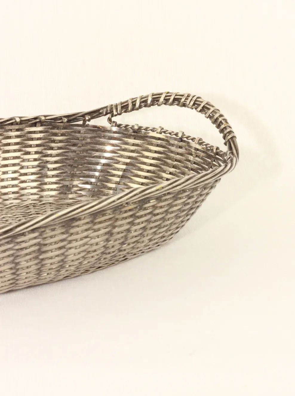 A close-up of a silver-colored brass basket with a curved handle. The hand-woven Woven Basket from Boga Avante Shop is laid on a neutral white background, showcasing its intricate geometric pattern and oval shape.