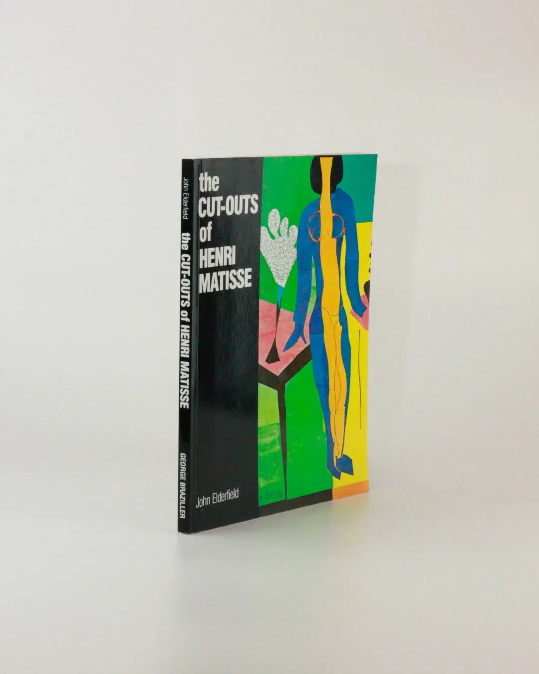 A book titled "The Cut Outs of: Henri Matisse" from Boga Avante Shop, authored by John Elderfield. Its cover displays an abstract artwork rich in bright colors and simplified human figures, characteristic of Matisse's mature cut-outs. The book stands upright against a plain background.