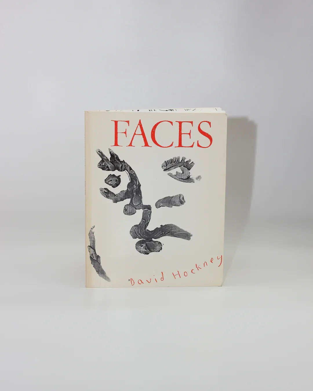 The "Faces" portrait art book from Boga Avante Shop displays abstract black and white facial features with bold red text on its cover. "David Hockney," the renowned artist, has his name elegantly scripted in red at the bottom. The book is shown upright against a plain gray background.