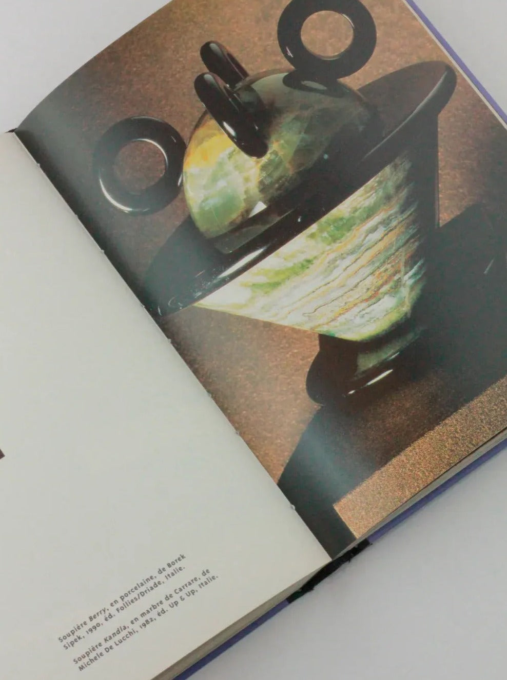 An open Objets Affectifs: Le Nouveau Design de la Table from Boga Avante Shop features a photo of an art piece consisting of a green and black vase with a lid and decorative rings. The opposite page contains text in Spanish, partially readable, and a small cropped image. The background is a neutral color, highlighting the diseño moderno of the book.