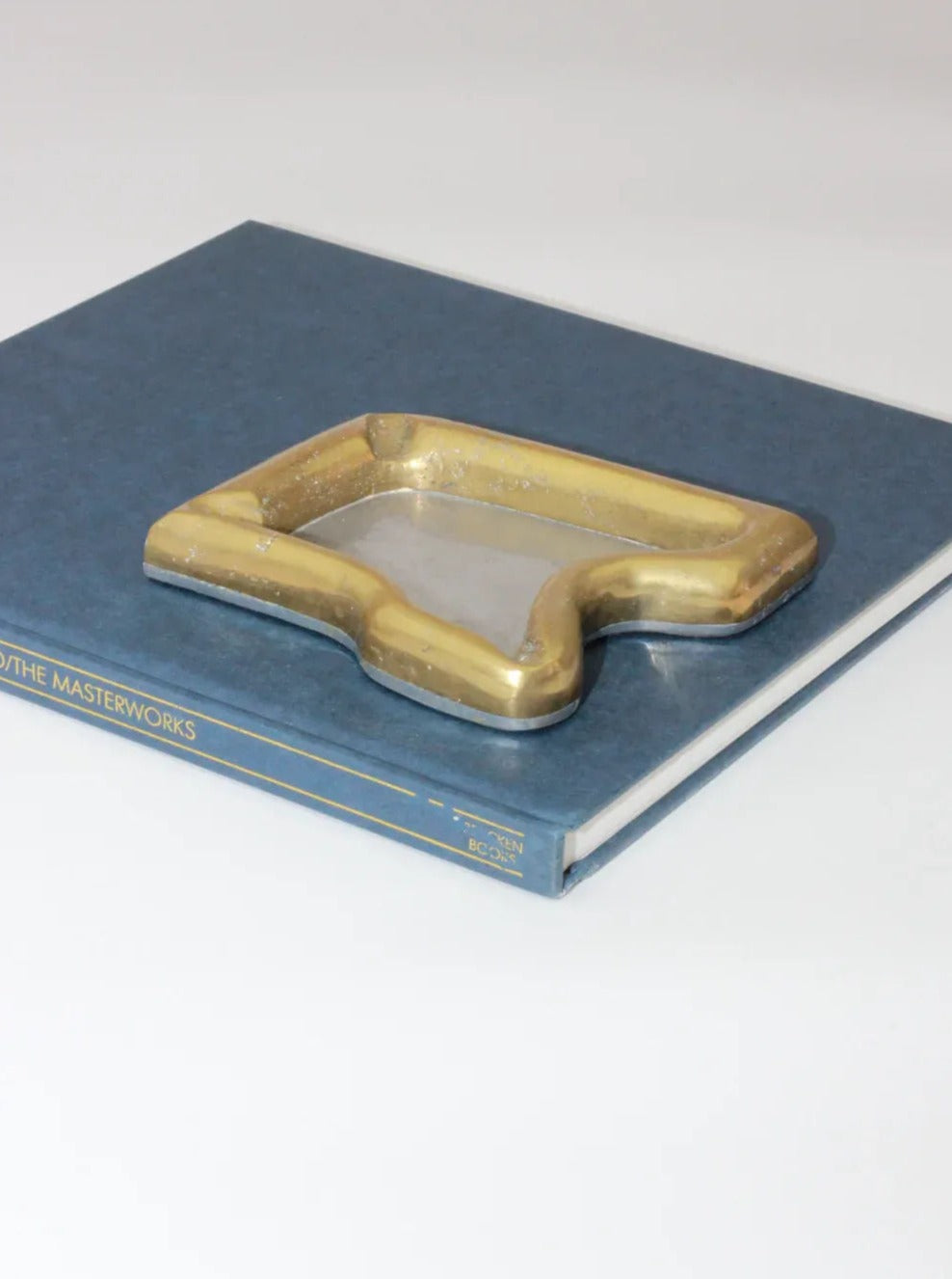 A Brutalist Ashtray from Boga Avante Shop rests on a closed blue book with the word "MASTERWORKS" partially visible on the cover. The background is a plain white surface, giving it a minimalist, almost Brutalist style aesthetic.
